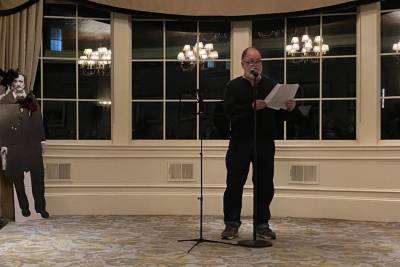 A bearded man in black sweater and black pants stands near a cardboard cutout of Edgar Allan Poe, reading a poem from a sheet of paper.