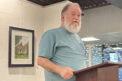 A white-bearded man in a green tee-shirt speaks from a podium in a library room.